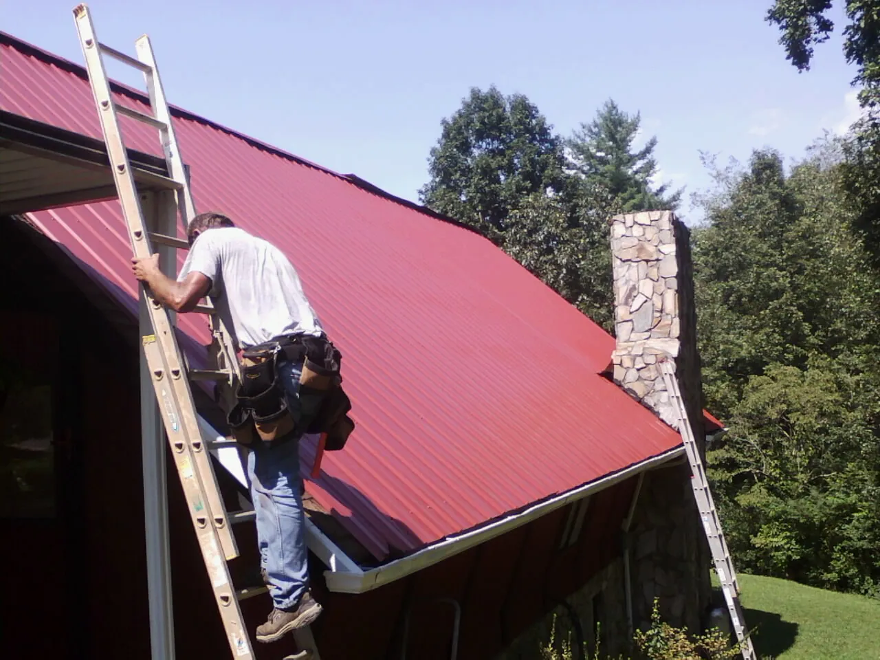 Roofer on a ladder beside a red metal roof and stone chimney — Soffit & Fascia Repair by Roofing Brothers in Del Valle